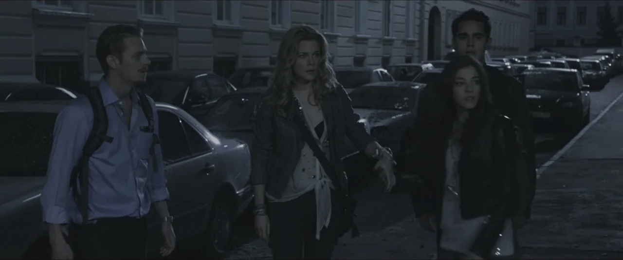 Darkest Hour (2017) movie still: fullbody — A quiet urban street at night is lined with numerous parked cars. Four young adults walk together along…
