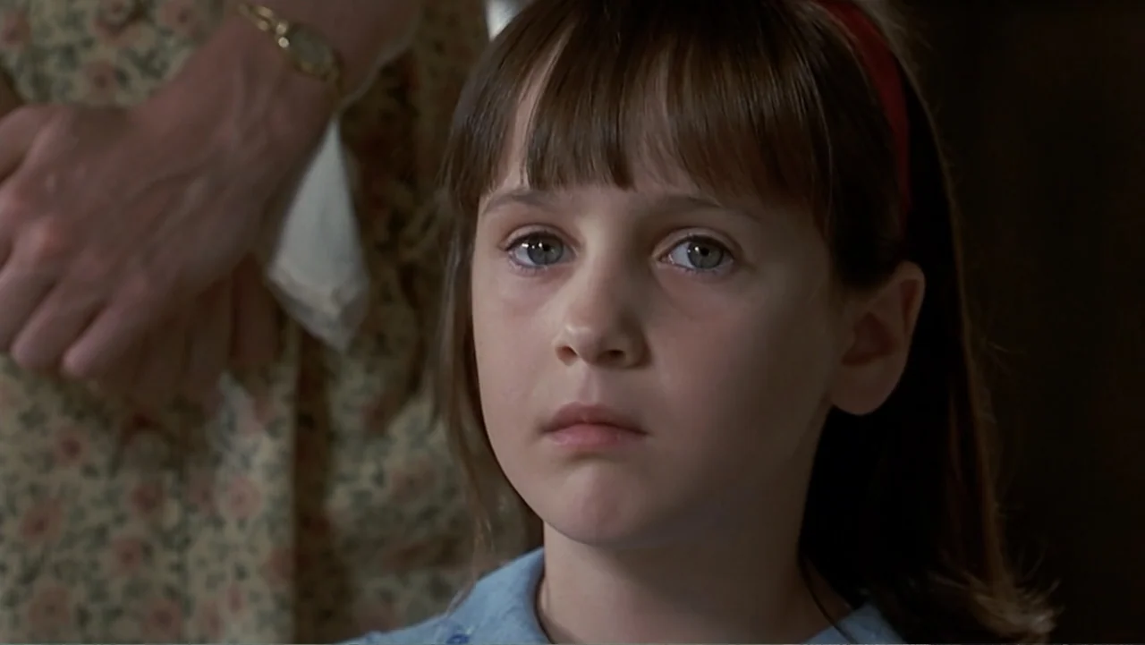 Matilda (1996) movie still: closeup — A close up of a young girl in an indoor setting. The girl looks forward with a steady gaze while wearing a red…