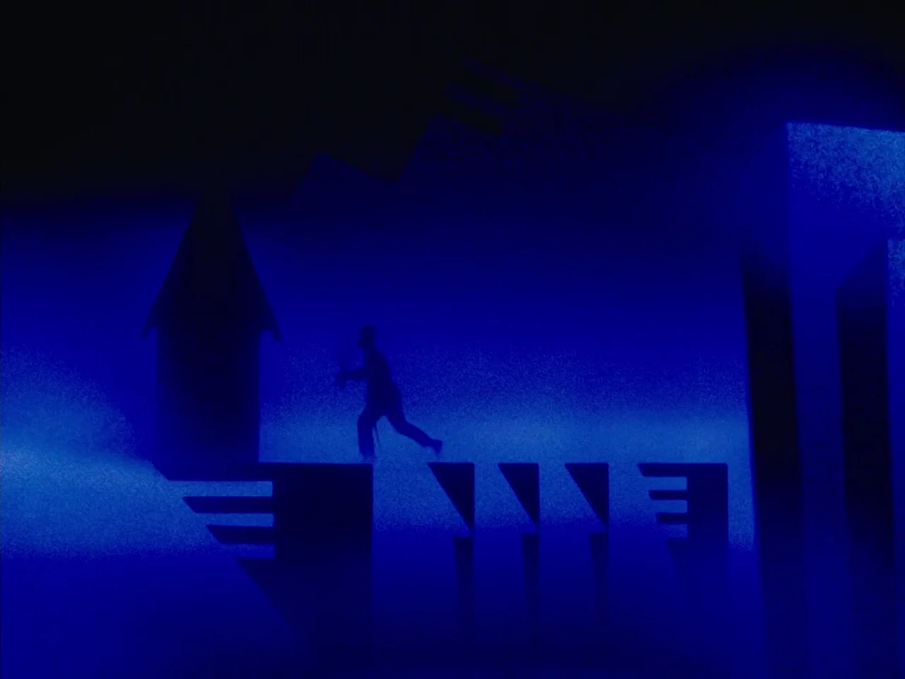 The Twentieth Century (2019) movie still: wide — A flat-topped urban structure at night is illuminated in deep, vibrant blue light. A silhouetted…