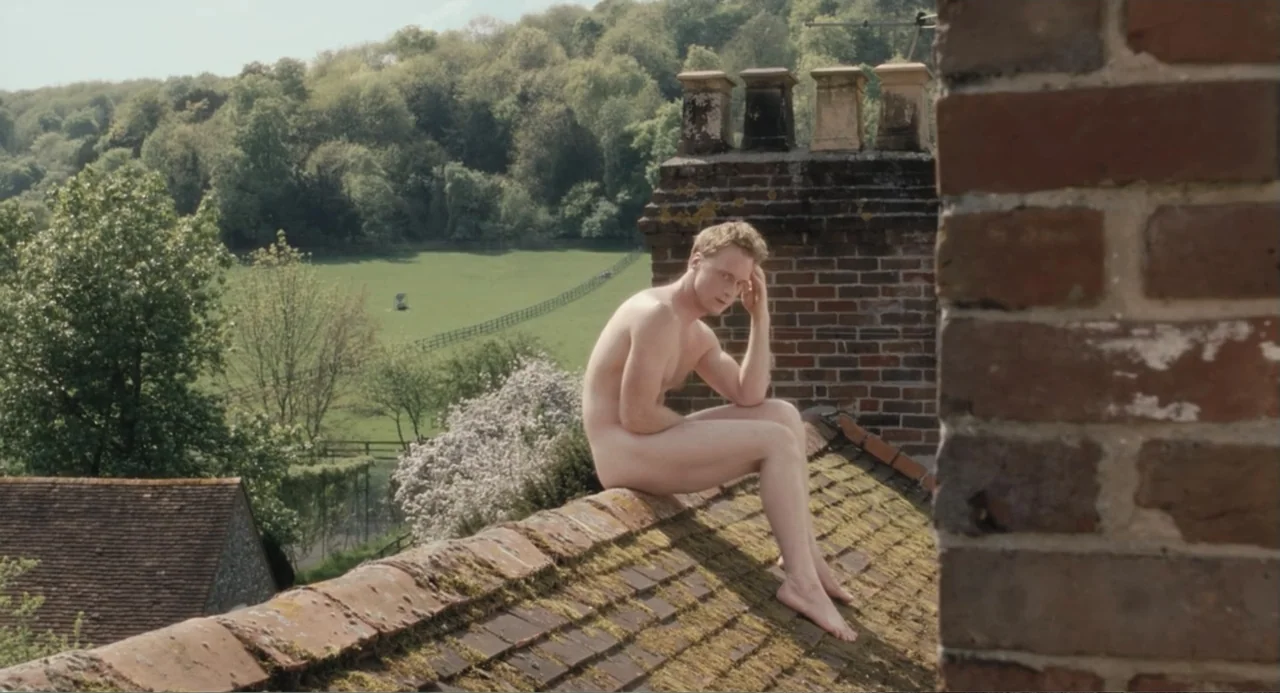 Death at a Funeral (2010) movie still: fullbody — A nude man sits atop a tiled roof overlooking a green rural valley. The setting includes brick…