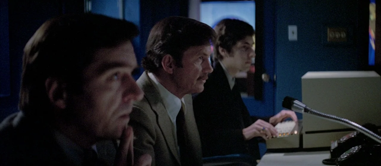 Three Days of the Condor (1975) movie still: medium — A dimly lit industrial control room features rows of vintage computing equipment and blue…
