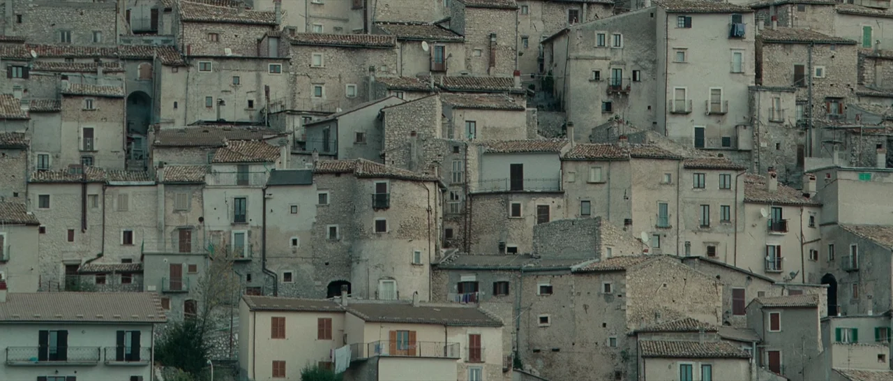 The American (2010) movie still: establishing — A dense cluster of old stone hillside houses fills the entire frame. Many buildings feature small…