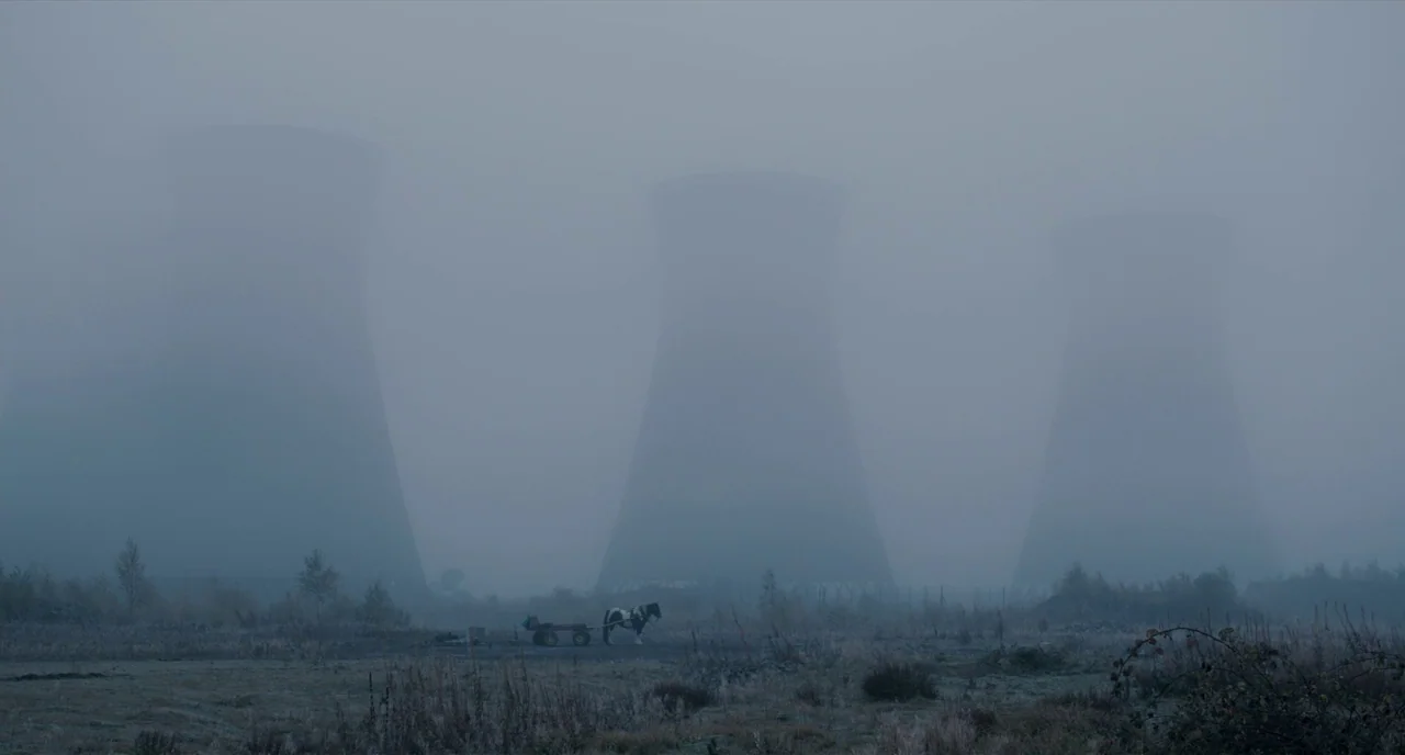 The Selfish Giant (2013) movie still: establishing — A field covered in thin mist features three large cooling towers in the distance. A lone horse…