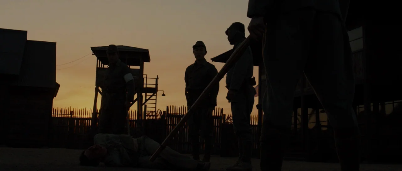 Unbroken (2014) movie still: wide — An outdoor military camp at dusk features a watchtower and wooden fence fortifications. Three soldiers in…