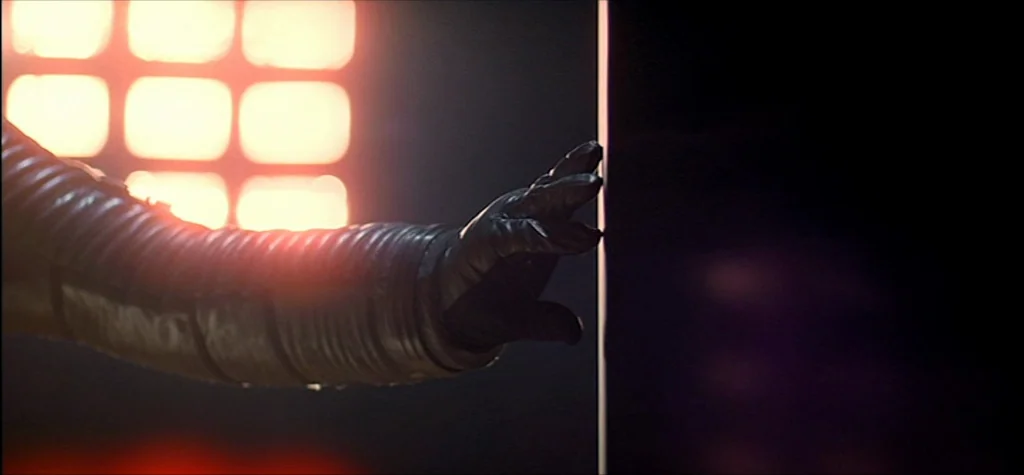 2001: A Space Odyssey (1968) movie still: closeup — A gloved hand in a ribbed sleeve reaches toward the edge of a black structure. Bright square…