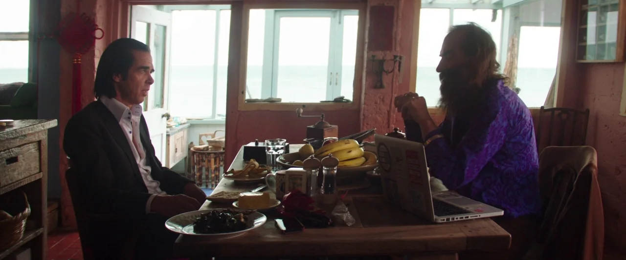 20,000 Days on Earth (2014) movie still: wide — A rustic sunroom features pink walls and large windows overlooking the ocean. Two men sit at a…
