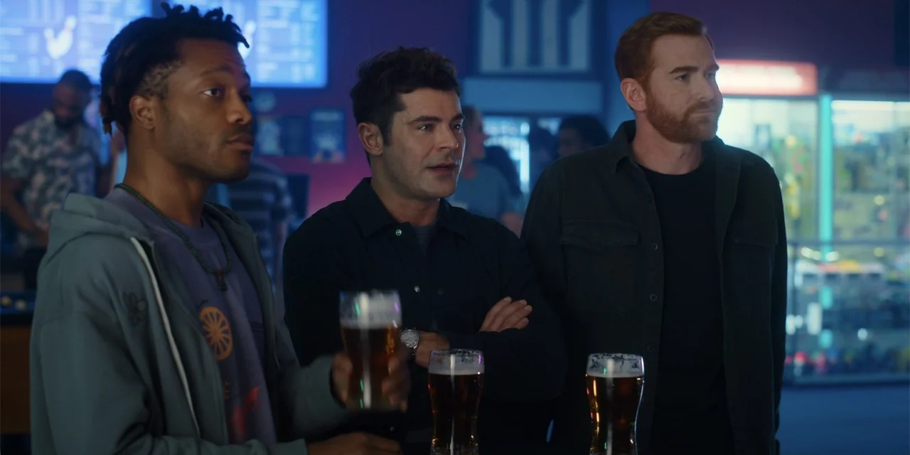 Ricky Stanicky (2024) movie still: medium — A dimly lit commercial recreation space features a counter and glowing digital displays. Three men stand…