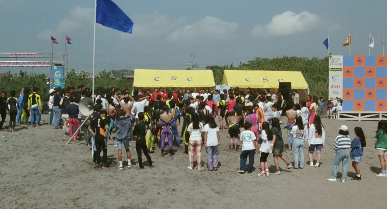 A Scene at the Sea (1991) movie still: wide — A crowded sandy beach hosts a surfing competition with two yellow canvas tents. Many people in…