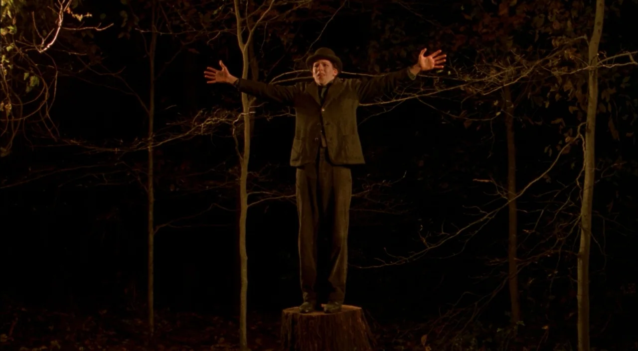 The Village (2004) movie still: fullbody — A dark, wooded area at night features a man standing on a tree stump. The man wears a suit and bowler hat…