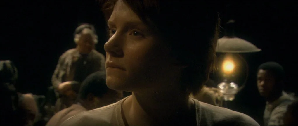 Manderlay (2005) movie still: closeup — A dark, dimly lit room filled with several people in the background. A woman in the foreground looks…