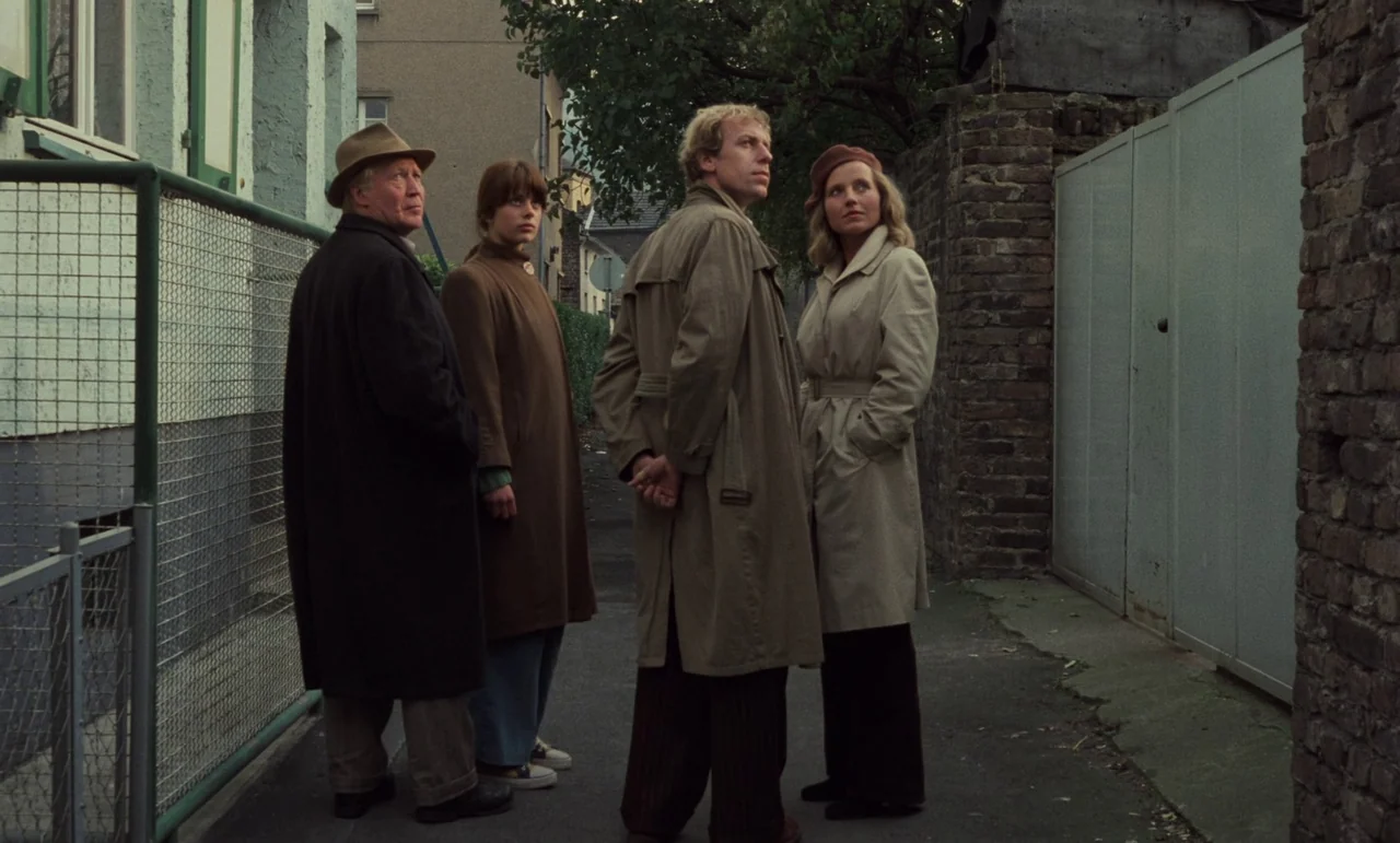 Wrong Move (1975) movie still: fullbody — A narrow paved alleyway is bordered by a brick wall and a wire fence. Four people stand in a line looking…
