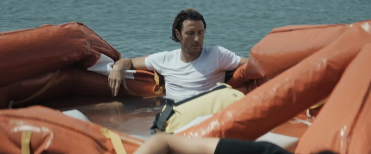 Great White (1998) movie still: medium — A man sits inside a bright orange rubber life raft floating on calm water. The man wears a white t-shirt…