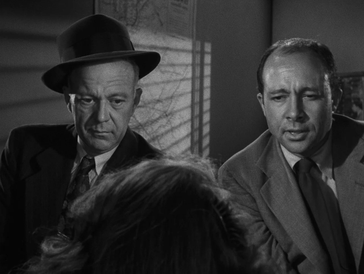 Pickup on South Street (1953) movie still: medium — An interior office with light streaming through venetian blinds on the wall. Two men in suits…