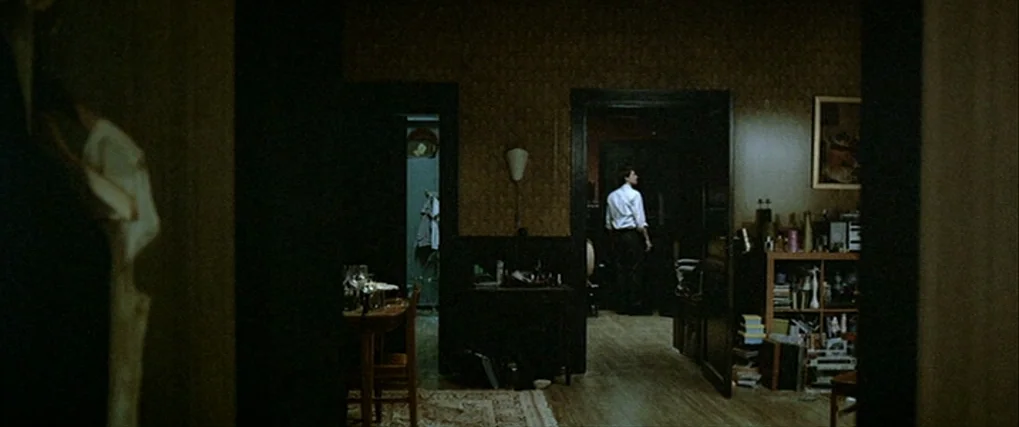 Next Door (2005) movie still: wide — An apartment interior features dark walls, wood flooring, and shelves cluttered with various items. A man in a…