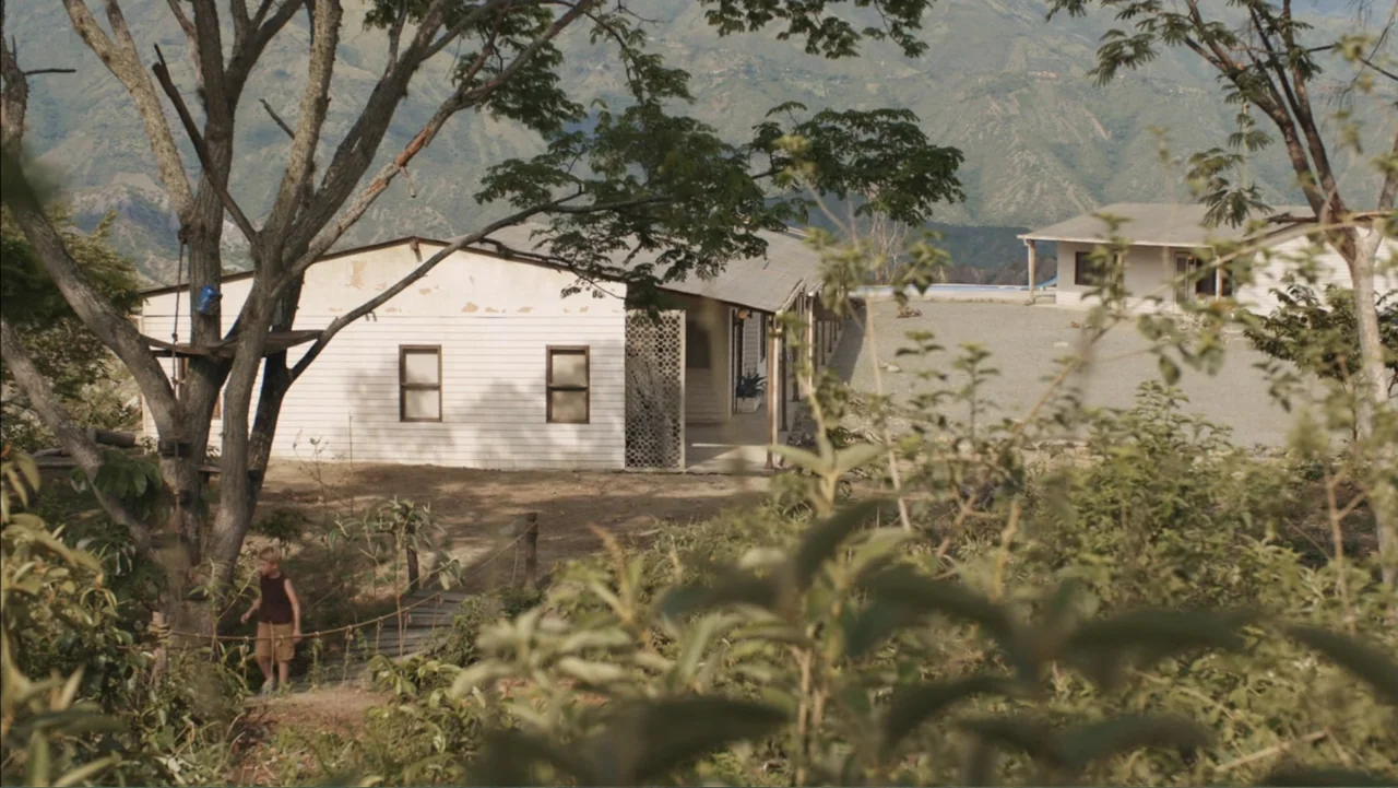 Boy (2010) movie still: wide — A rural landscape features white wooden buildings set against a backdrop of distant mountains. A young child walks…