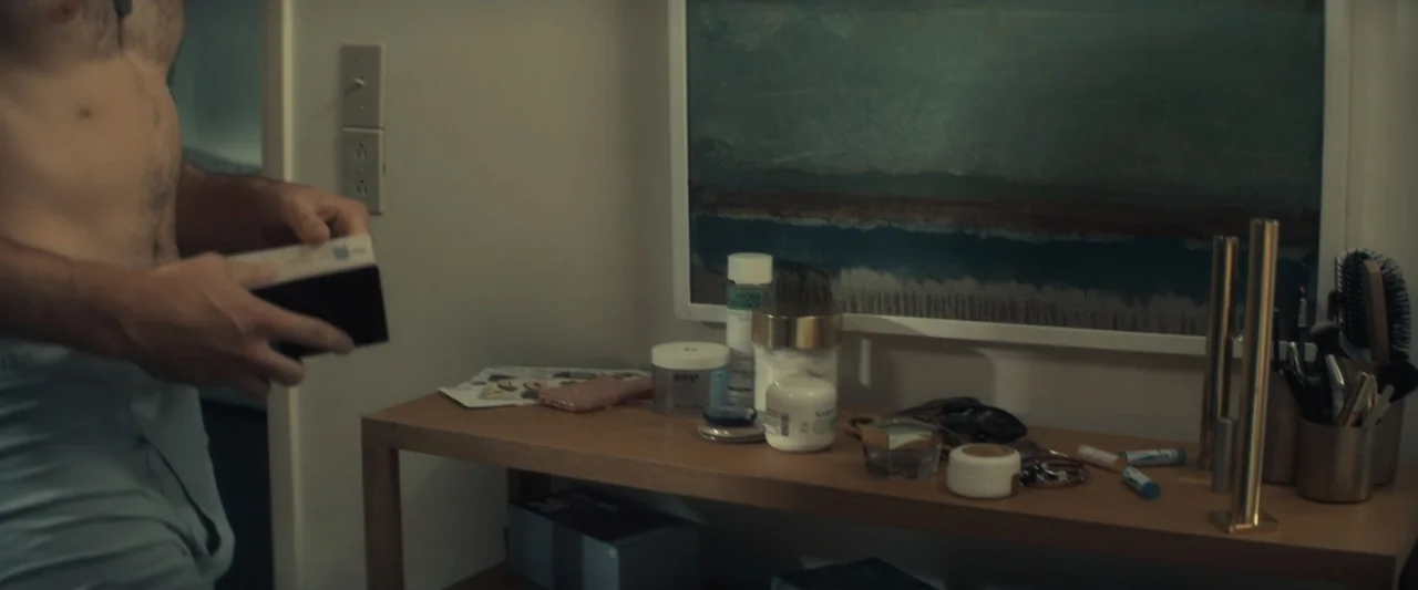 I Love America (2022) movie still: medium — A modern bedroom contains a wooden vanity cluttered with various skincare jars and bottles. A shirtless…