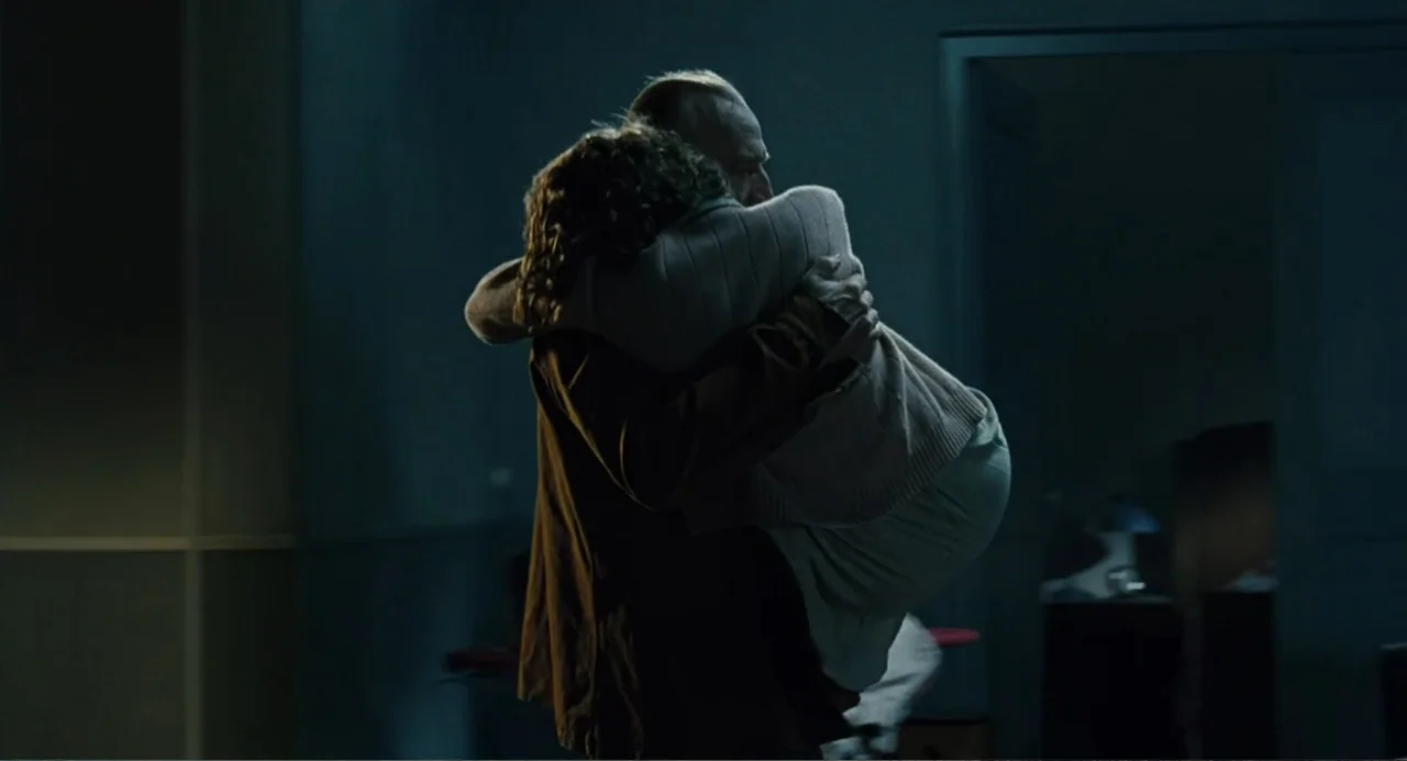 Le Havre (2011) movie still: medium — A dimly lit room with cool shadows and minimal background detail. A man holds a woman in his arms during an…