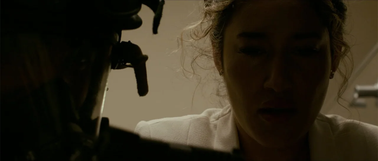 The Vault (2017) movie still: closeup — A dimly lit interior room features a close perspective of a woman and a tactical figure. The woman maintains…