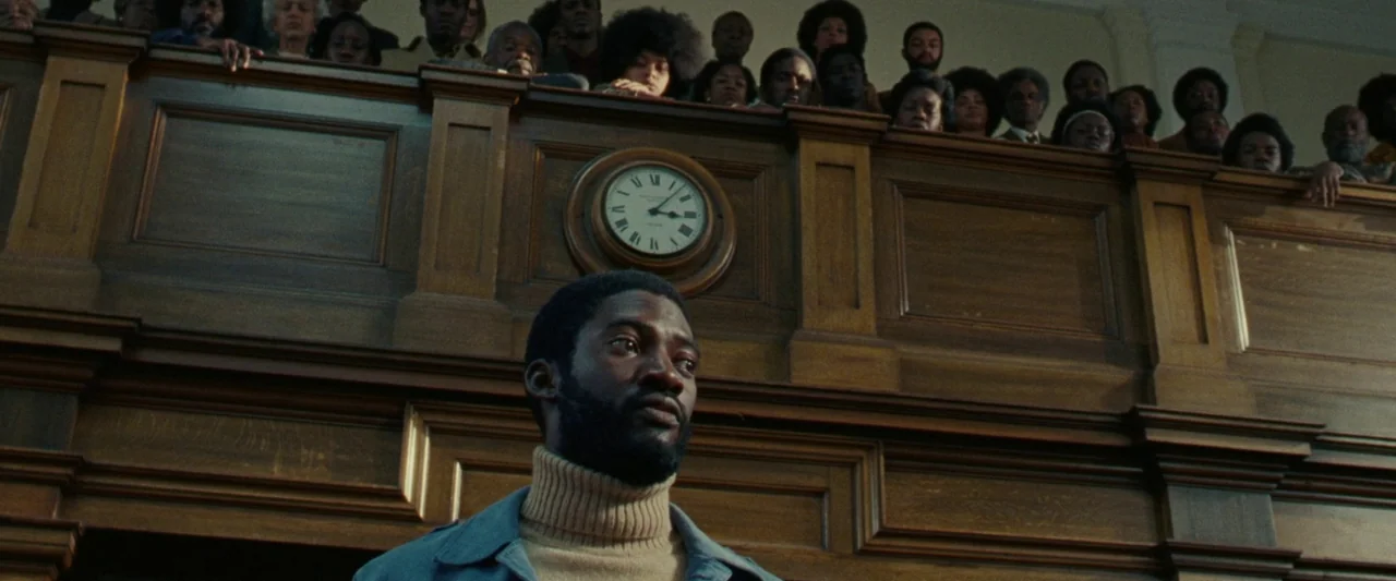 Mangrove (2020) movie still: medium — A dark wood-paneled wall features a circular analog clock mounted above the subject. A large group of diverse…
