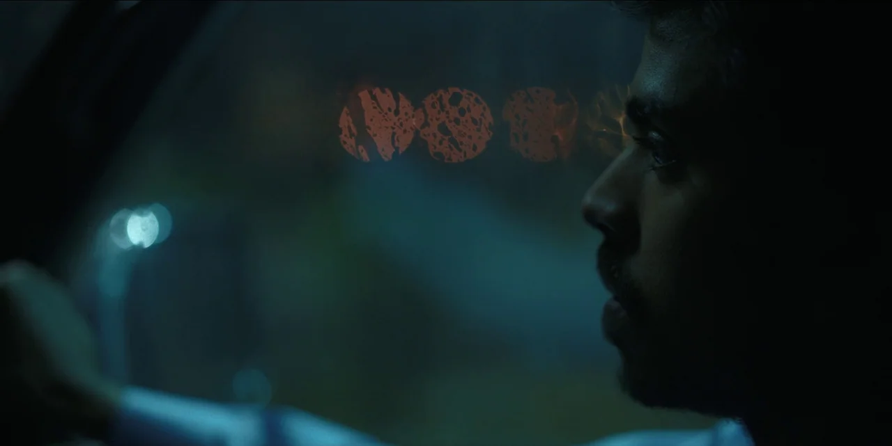 The White Tiger (2021) movie still: closeup — A man drives a car at night while rain beads on the window. Outside, city lights create blurred,…