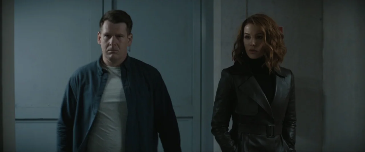Canary Black (2024) movie still: medium — A dimly lit industrial hallway features plain grey walls and a metallic double door. A man in a blue shirt…