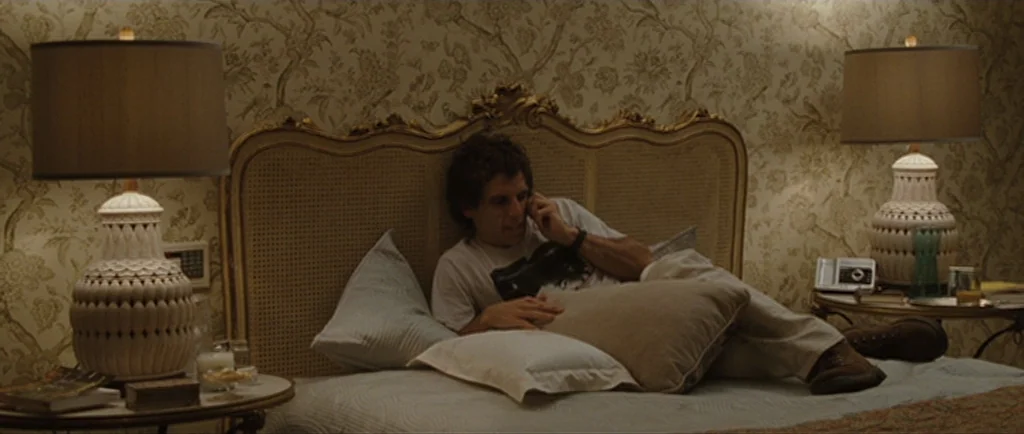 Greenberg (2010) movie still: medium — A hotel room features ornate floral patterned wallpaper and a large tufted headboard. A man lies across the…