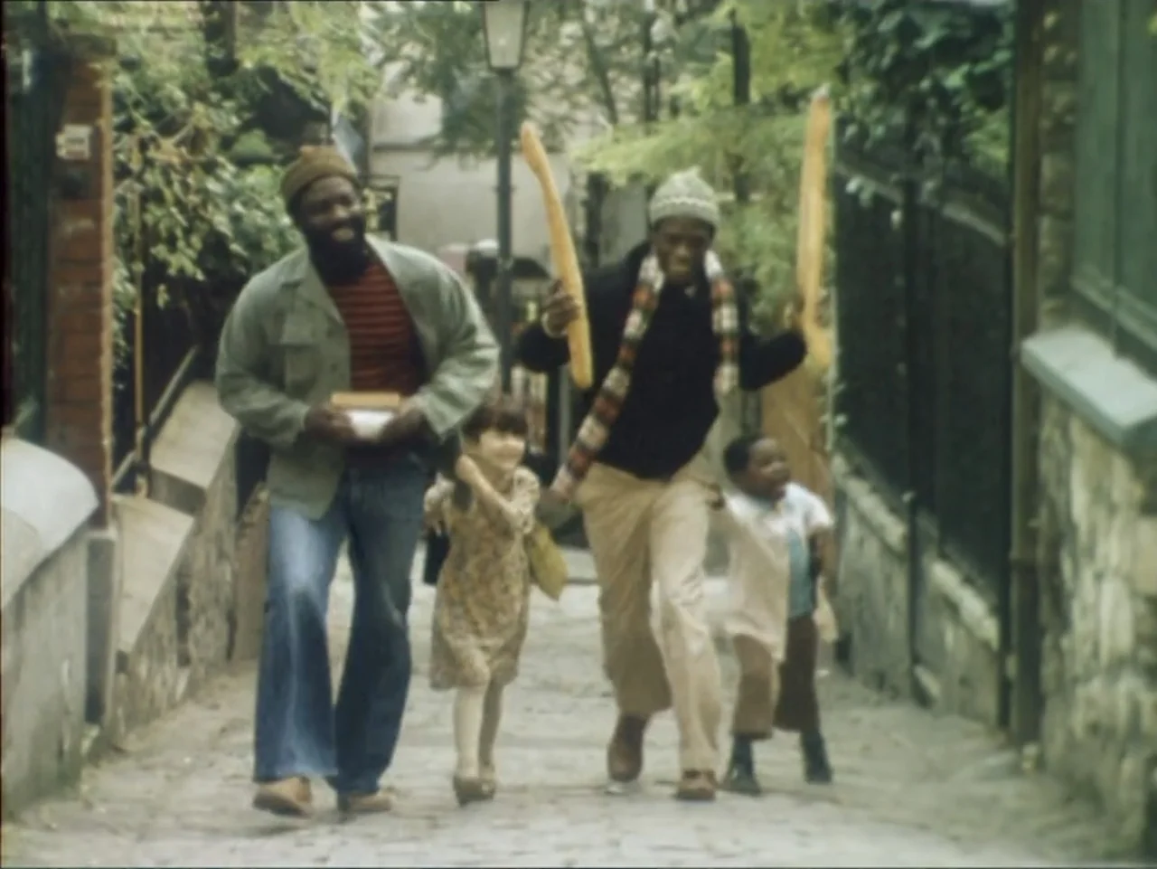 A Dessert for Constance (1981) movie still: fullbody — An outdoor cobblestone path flanked by stone walls and metal fencing appears in daylight. Two…