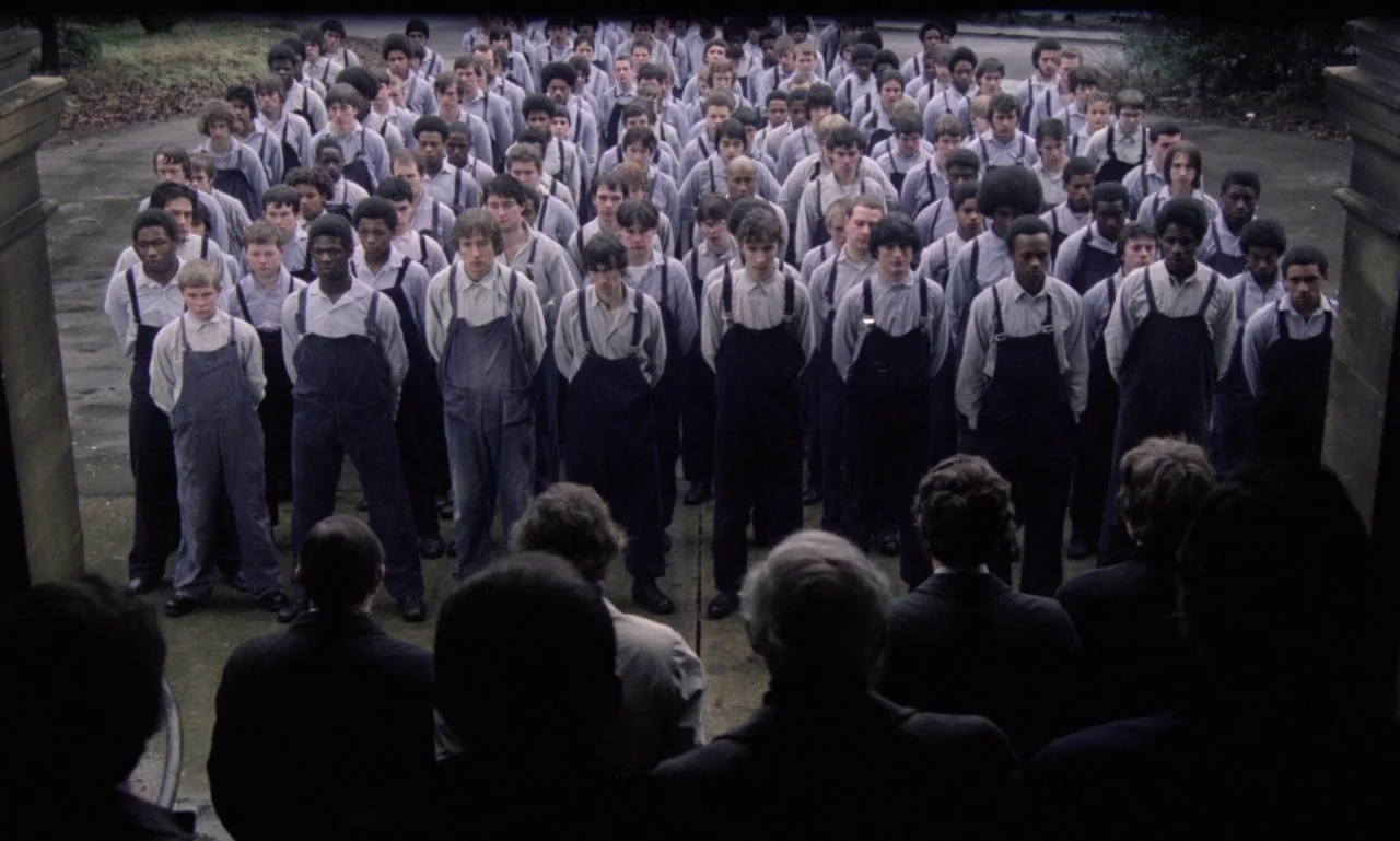 Scum (1979) movie still: wide — A vast crowd of young men in overalls stands in organized rows outdoors. Figures in suits observe the group from the…