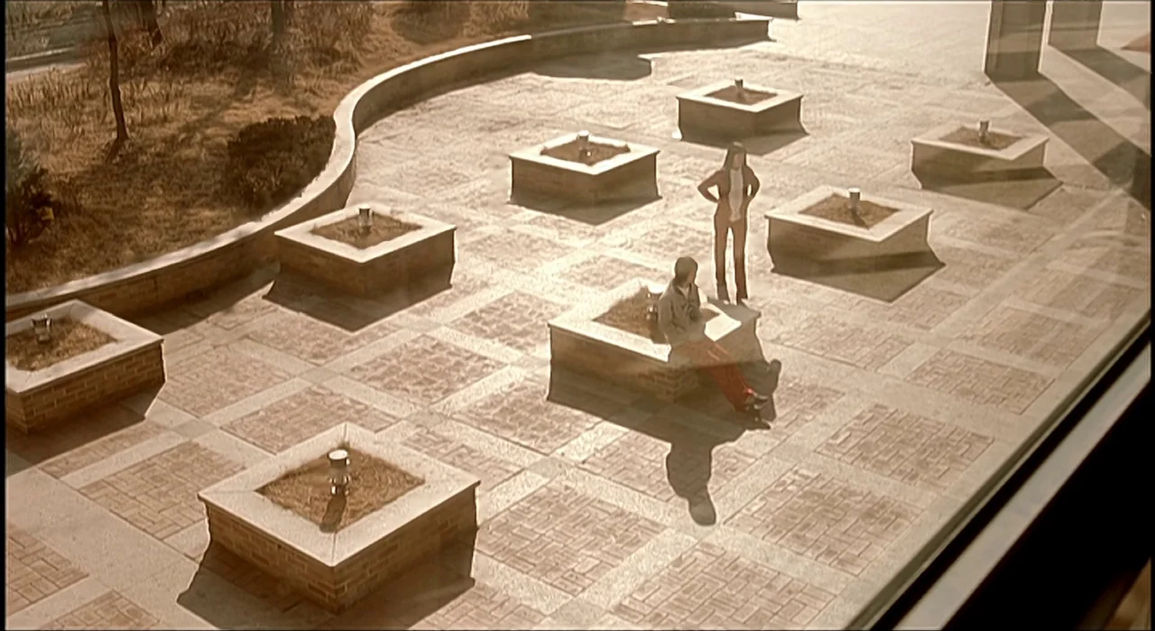 Voice (2005) movie still: wide — A paved outdoor plaza features numerous square stone planters arranged in a repeating grid pattern. A man sits on…