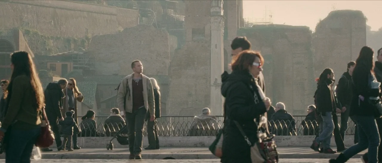Spring (2014) movie still: fullbody — A crowded pedestrian bridge overlooks ancient Roman ruins bathed in golden afternoon sunlight. Numerous people…