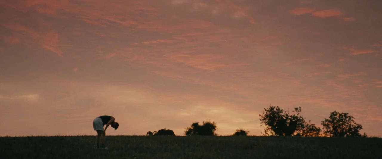 The Imitation Game (2014) movie still: wide — An expansive open grassy field under a vibrant orange and pink sunset sky. A young man stands alone in…