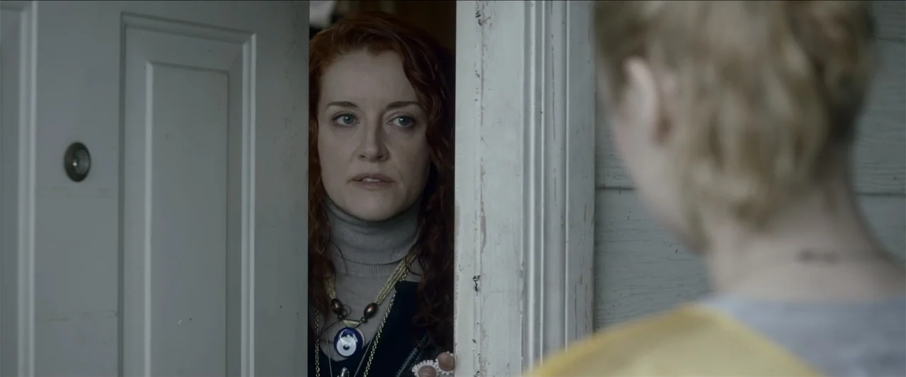 The Hollow Child (2018) movie still: medium — A woman looks through the narrow opening of a white wooden door frame. She is positioned inside a home…