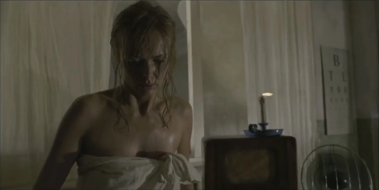 Exorcist: The Beginning (2004) movie still: medium — A dimly lit room contains a table with a burning candle and a fan. A woman with wet hair and a…