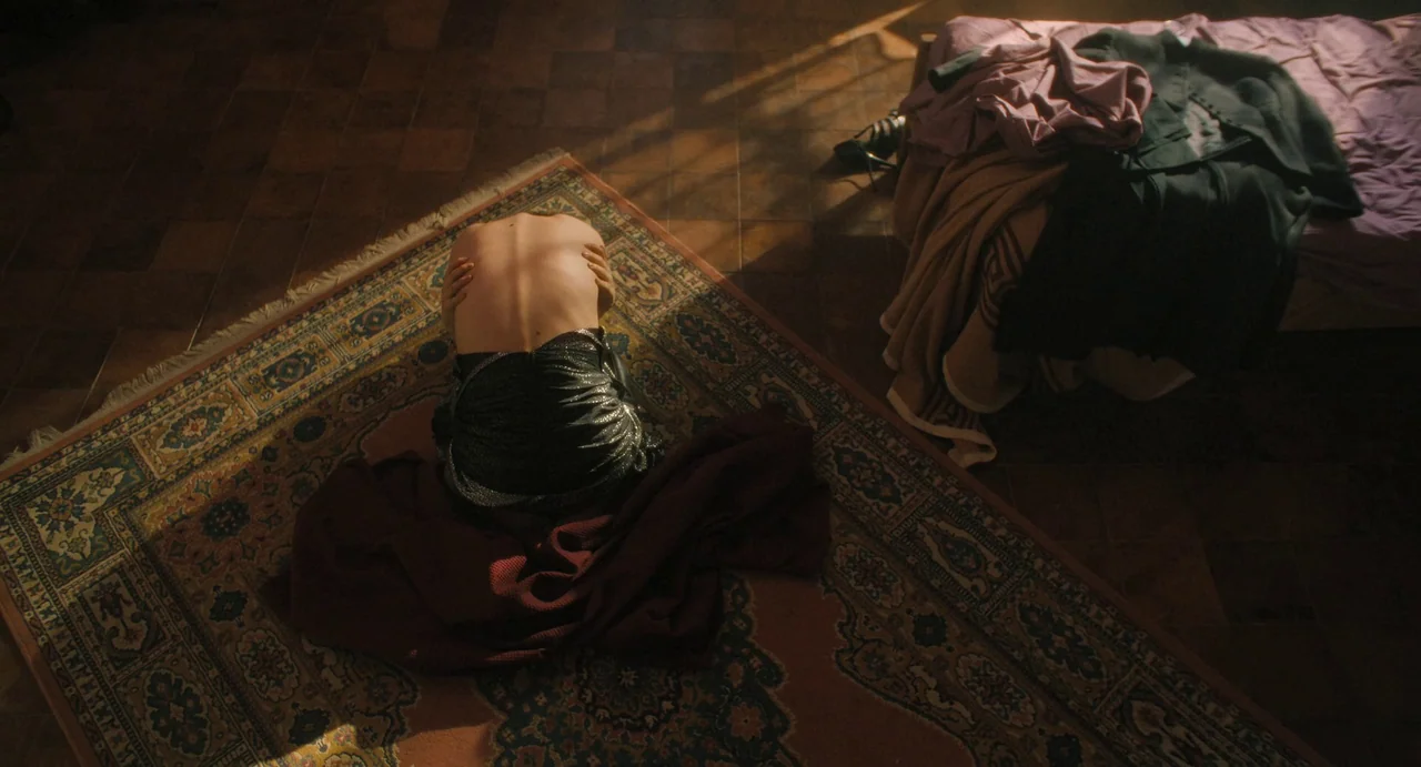 The First Omen (2024) movie still: wide — A tiled room with a patterned area rug and a bed covered in clothes. A person kneels on the rug, facing…