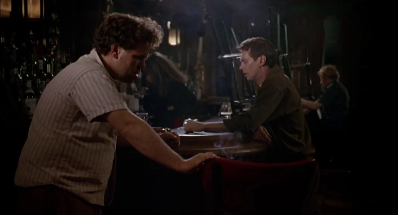 Trees Lounge (1996) movie still: medium — A dimly lit bar features dark wood paneling, rows of bottles, and overturned chairs. Two men stand at the…