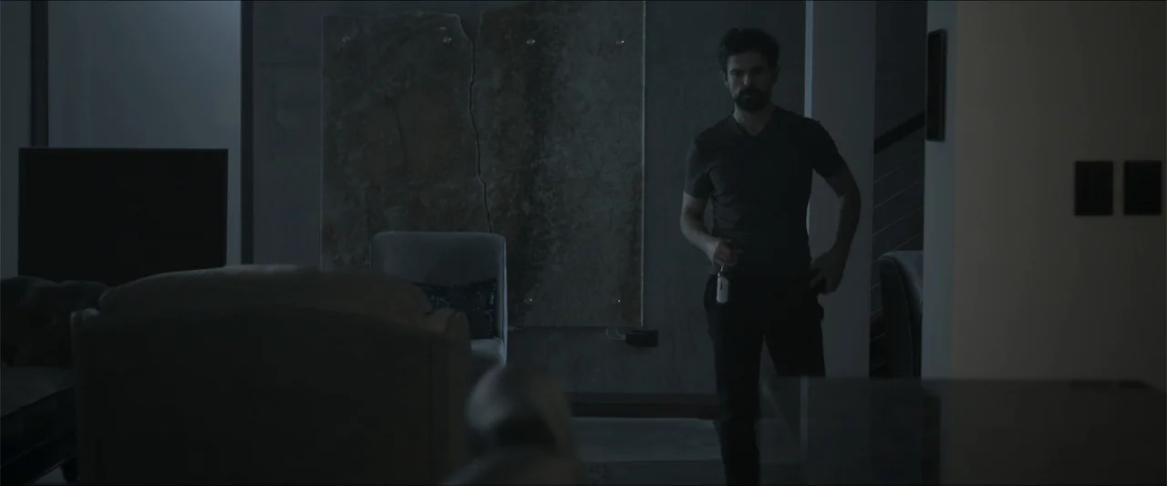 Los habitantes (2023) movie still: medium — A modern living room contains a large stone panel wall and dark furniture in dim lighting. A man with a…