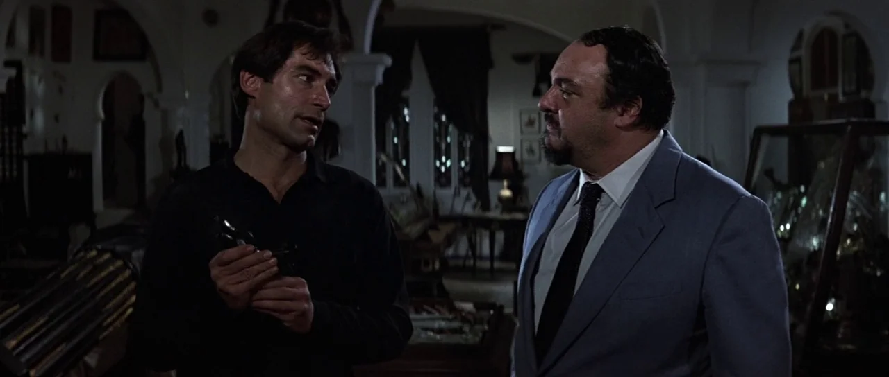 The Living Daylights (1987) movie still: medium — A dimly lit room filled with miscellaneous antiques and glassware on dark shelves. Two men in…