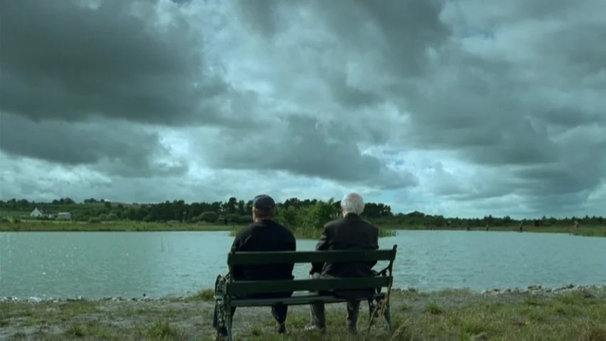 Garage (2007) movie still: wide — A quiet lake under a heavy, overcast sky stretches toward a distant green shoreline. Two men sit on a wooden bench…