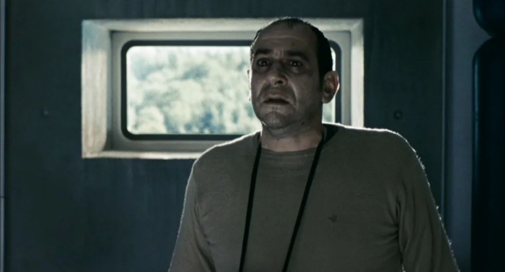 Timecrimes (2007) movie still: medium — A stark room features a rectangular window showing a blurred green exterior landscape. An adult man with…