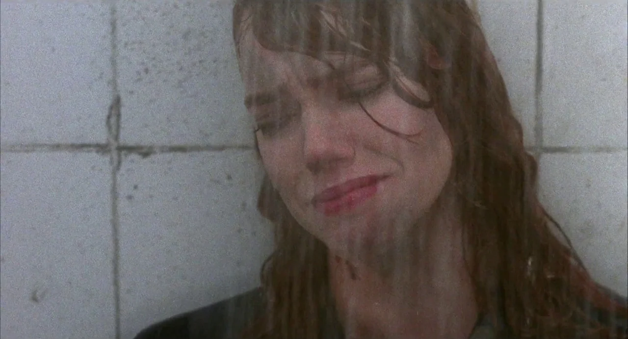 Hardware (1990) movie still: closeup — A woman stands in a shower with gray, tiled walls behind her. She keeps her eyes closed as water runs down…