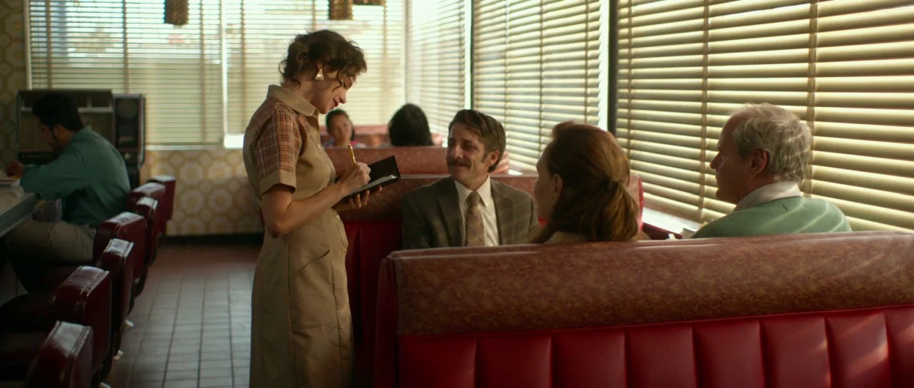 Faults (2014) movie still: medium — A 1970s diner with red vinyl booths and Venetian blinds covering large windows. A waitress stands at a table…