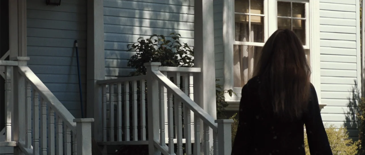Carrie (2013) movie still: medium — A light blue wooden house features a white porch railing and a small window. A person with long brown hair walks…