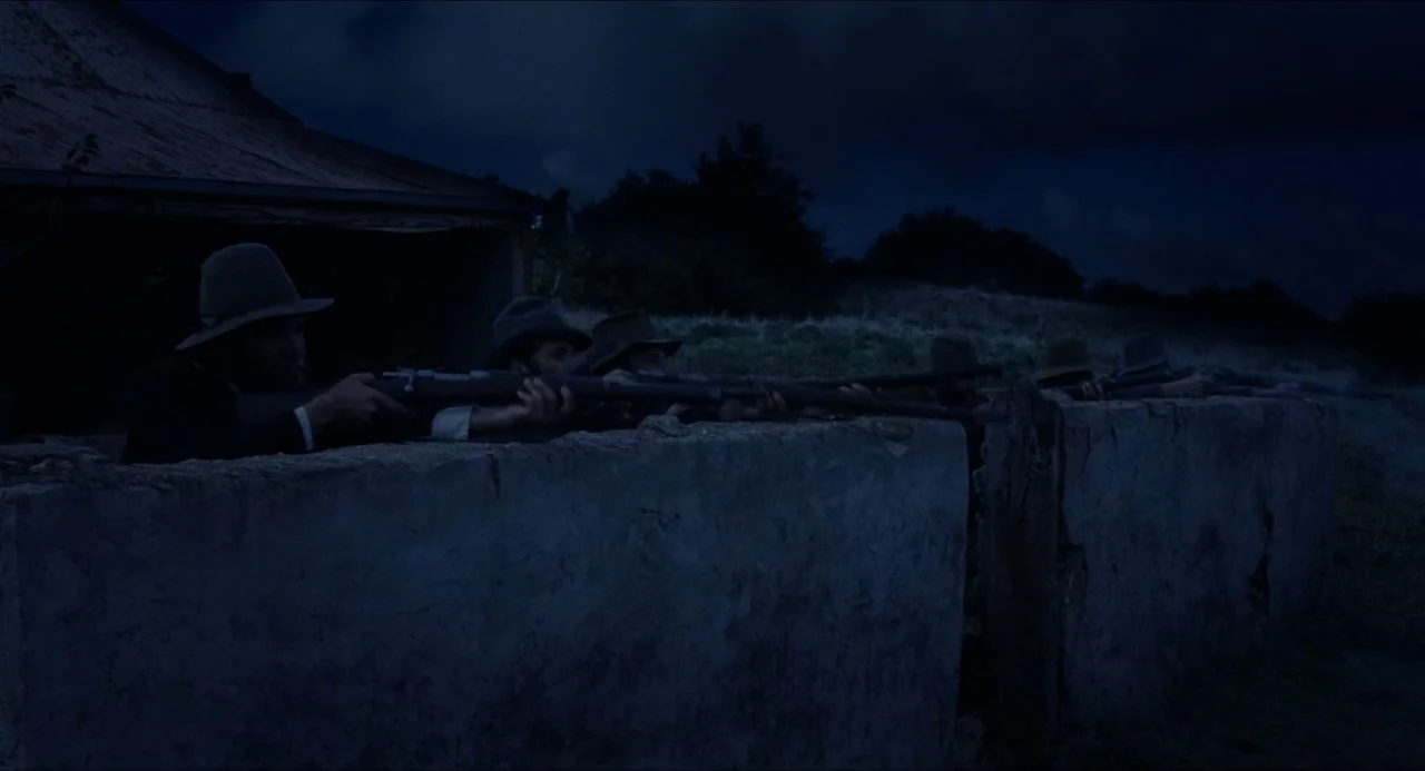 Breaker Morant (1980) movie still: wide — A group of armed men crouch behind a low stone wall at night. They aim rifles across the top of the wall…