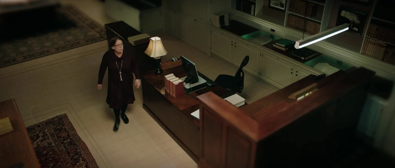 American Animals (2018) movie still: wide — A quiet, dimly lit archive room with wooden furniture and rows of books. A woman in a dark dress walks…