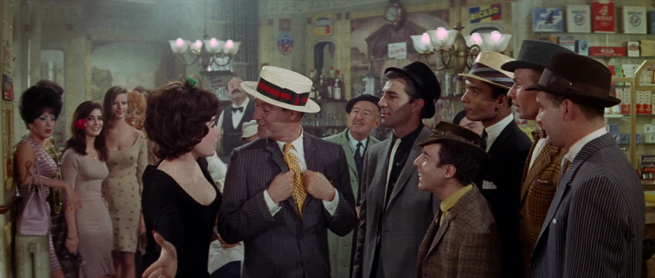 Irma La Douce (1963) movie still: medium — A crowded bar interior with shelves of tobacco products and alcohol bottles visible behind. A group of…