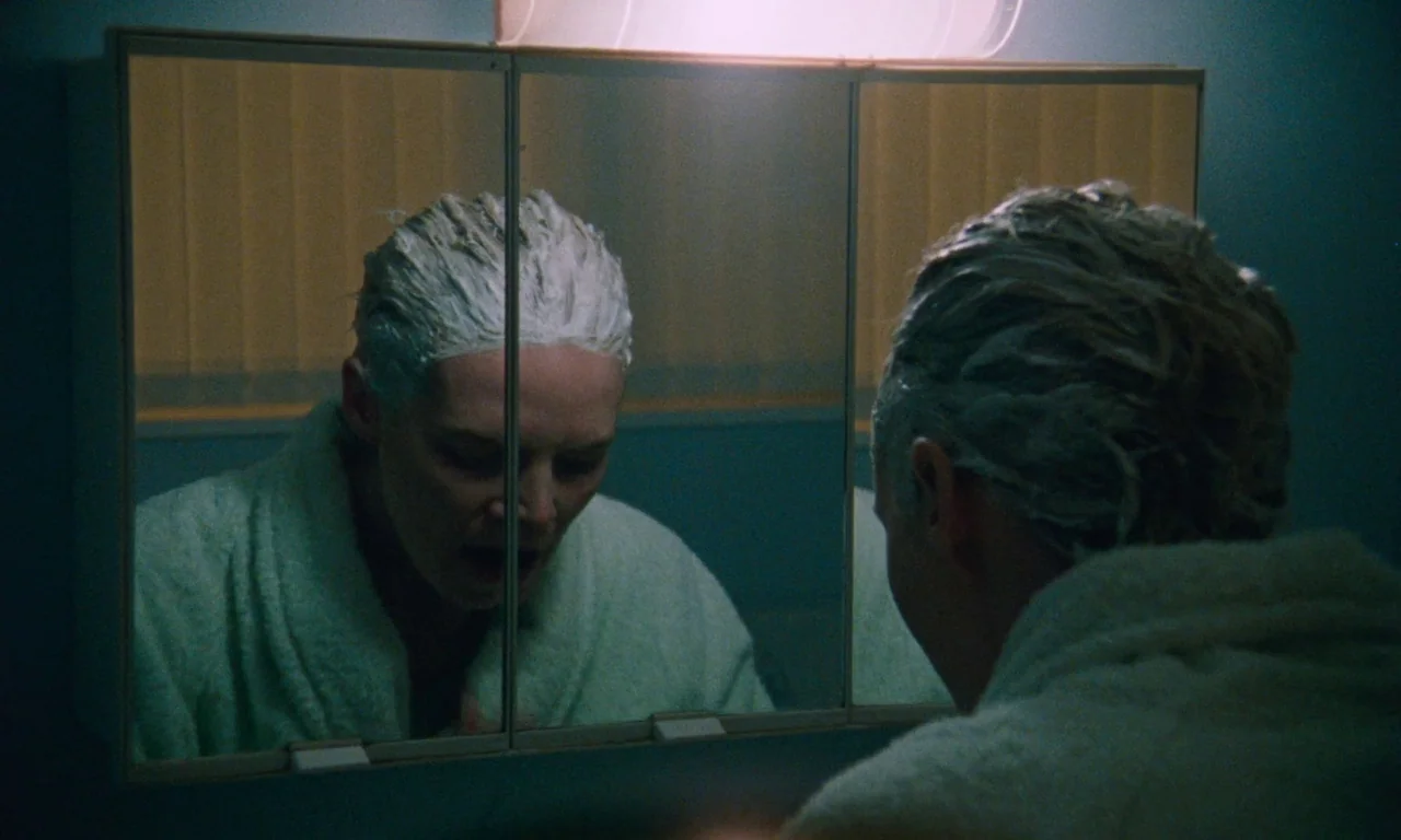 Blue Jean (2022) movie still: closeup — A woman stands in a dimly lit bathroom with walls painted a deep teal. She gazes at her own reflection in…
