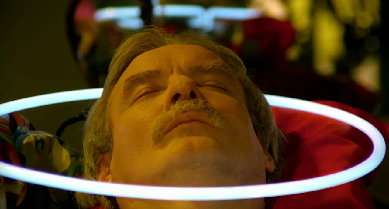 Liquid Sky (1982) movie still: closeup — A man lies with his eyes closed inside a hair salon under a hood. A bright circular white neon ring light…