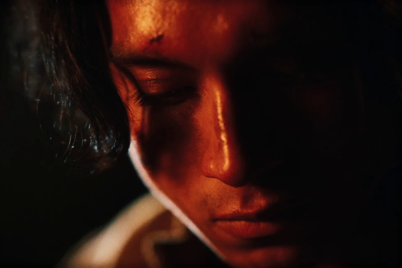 The Settlers (2023) movie still: closeup — A dark, minimally lit interior space frames a person in close proximity. The individual gazes downward…
