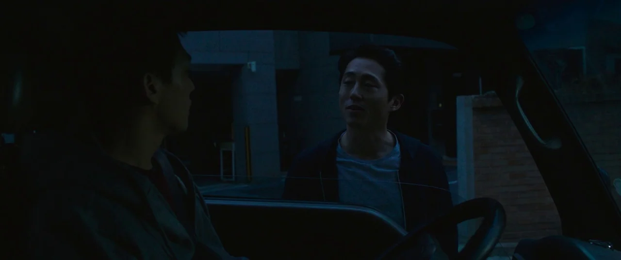 Burning (2018) movie still: medium — Two men converse through the open window of a vehicle parked on a city street. The man outside the car stands…