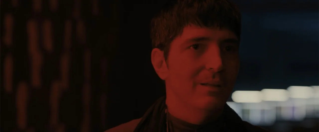 Afraid (2024) movie still: closeup — A man stands in a dark, out-of-focus indoor space with red light exposure. He is positioned slightly off-center…